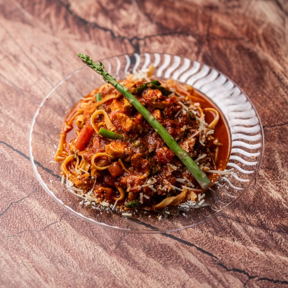 Pasta with red sauce, chicken, shredded cheese, and a green asparagus spear on a clear plate.